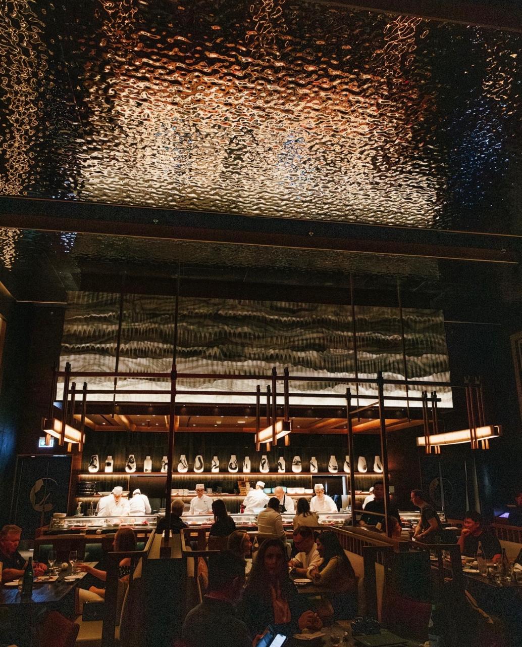 Dimly lit fine dining room with an open sushi counter, rippled metallic ceiling and guests at candlelit tables